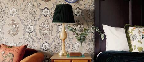 A bedroom with a patterned wallpaper, a wooden nightstand with drawers, a green lamp, a decorative plate, a dark headboard, and a plaid rug.
