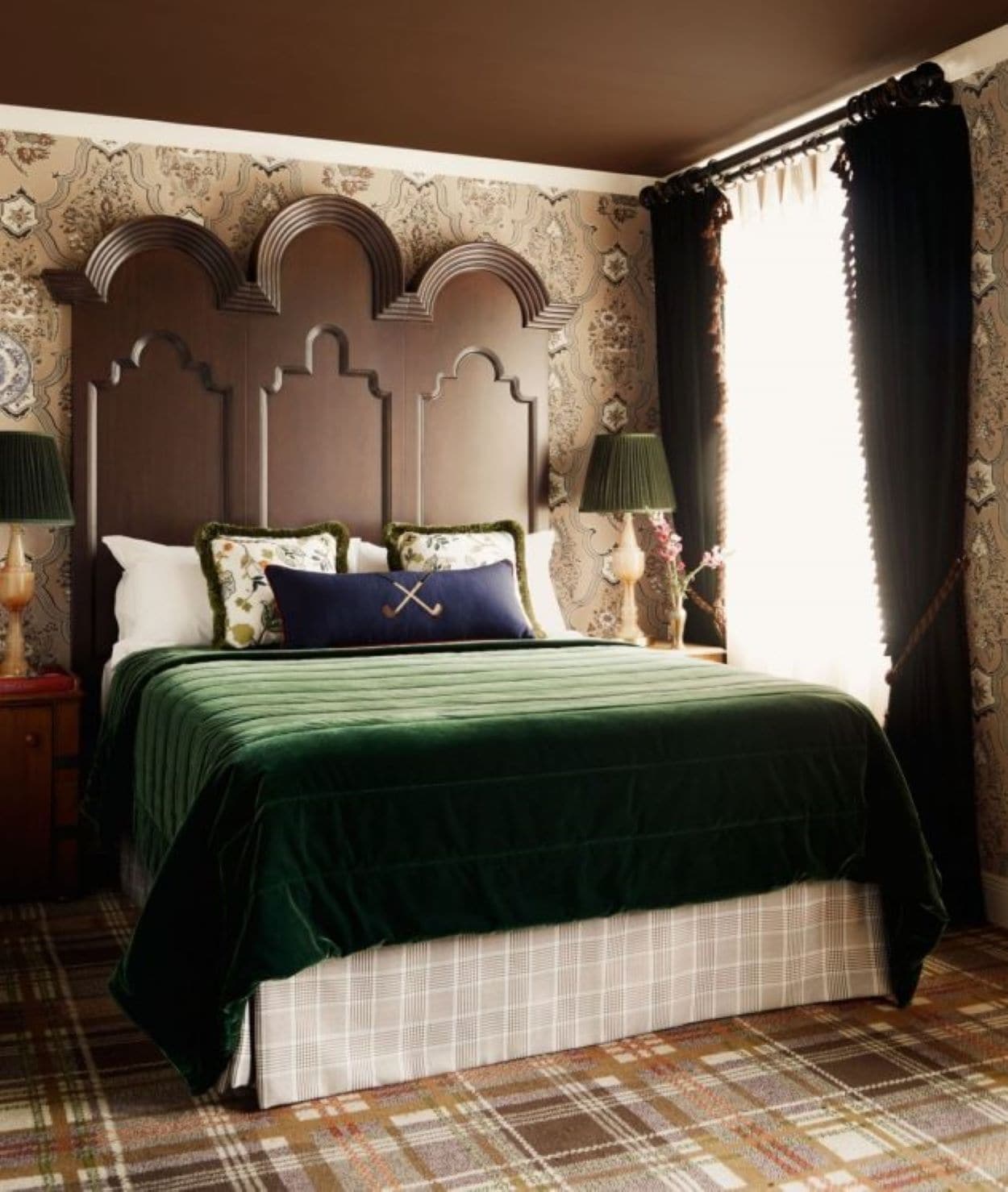 A bedroom with a large bed, a headboard with decorative arches, two bedside lamps, and a plaid-patterned rug.