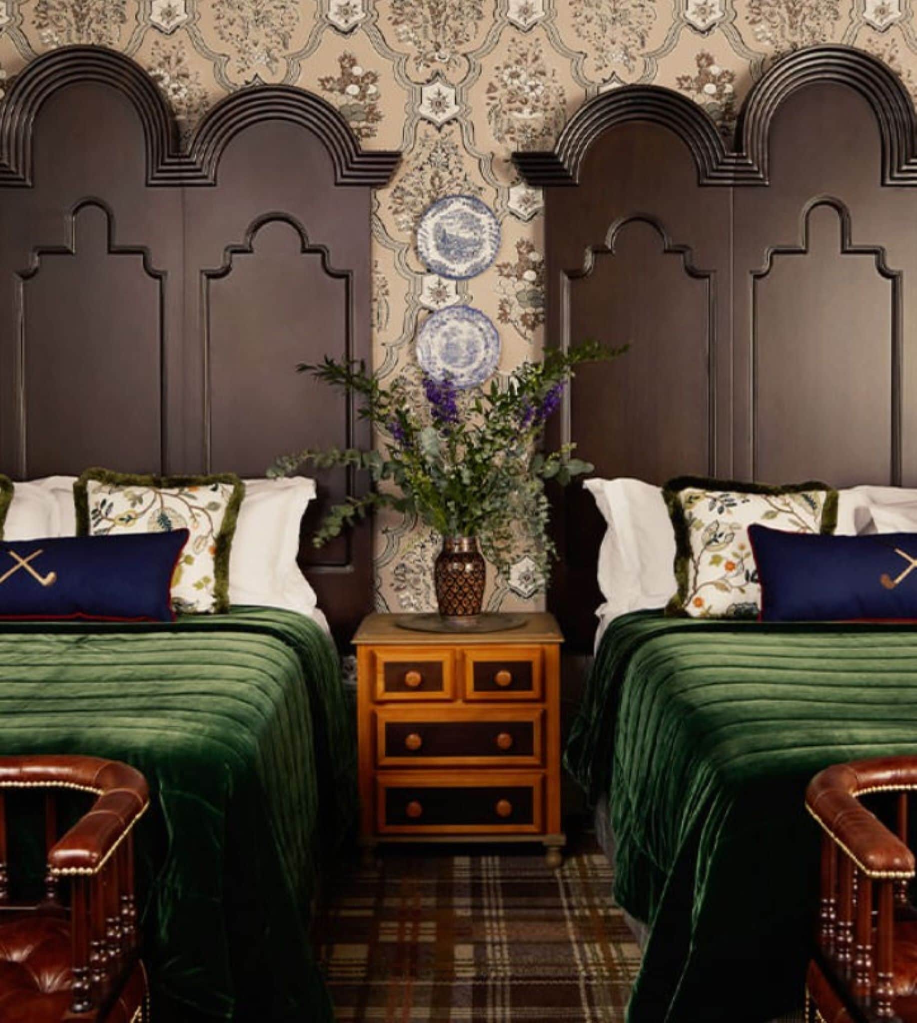 A room with two beds, a nightstand, a vase with flowers, and a patterned wallpaper.