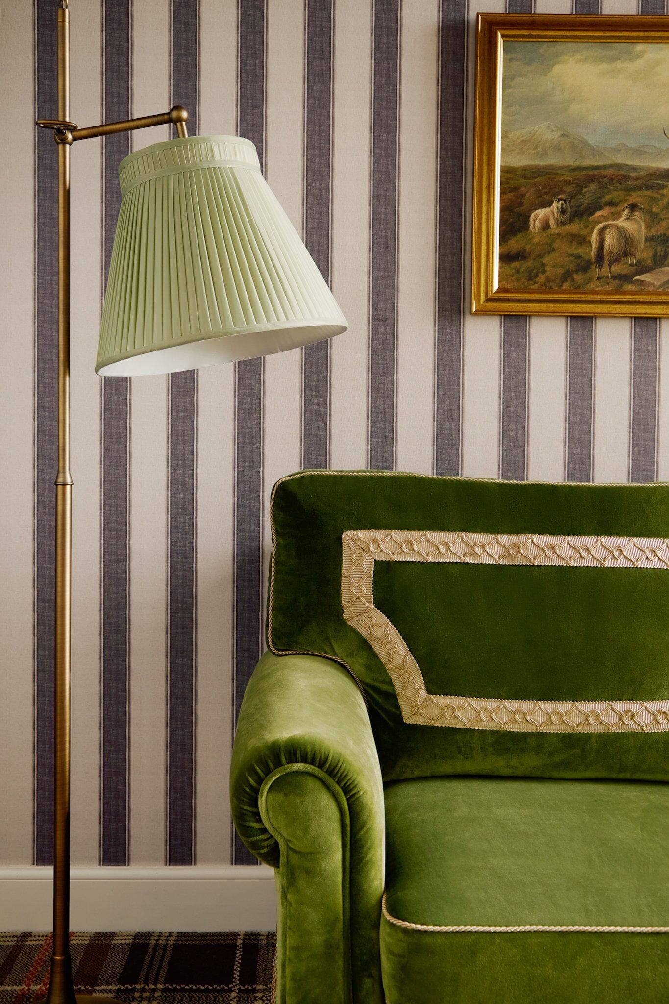 Un fauteuil vert recouvert de tissu, agrémenté d’un coussin à motifs, une lampe sur pied et un papier peint à rayures en arrière-plan.