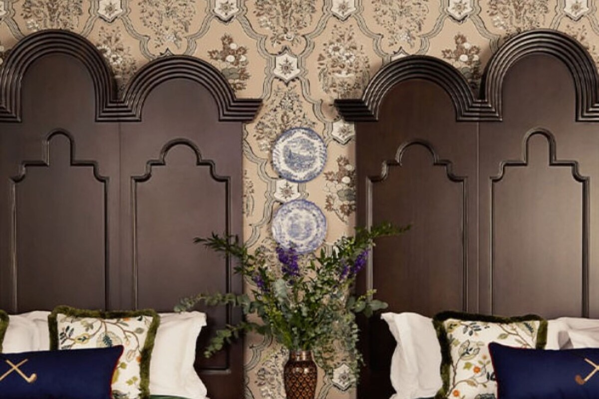 A room with two beds, a nightstand, a vase with flowers, and a patterned wallpaper.