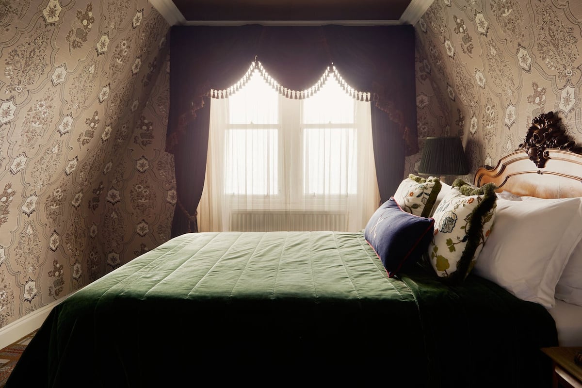 A bedroom with a green bedspread, floral patterned wallpaper, a window with sheer curtains, and a wooden headboard.