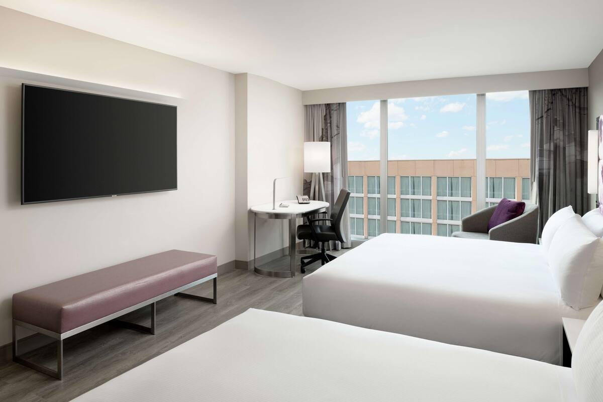 A hotel room with a large bed, a smaller bed, a desk with a chair, a bench, a TV, and a view of the city.