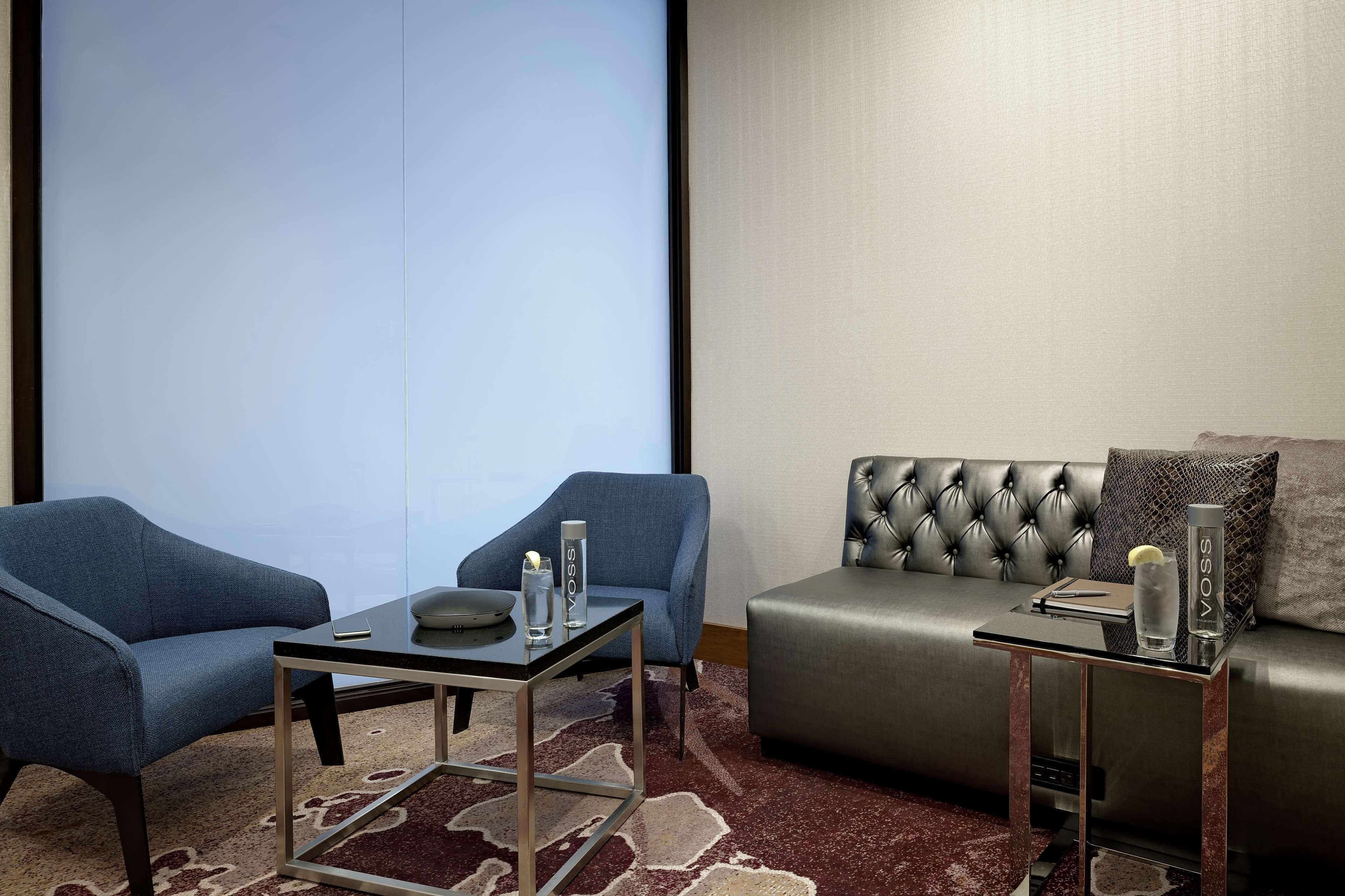 Ett modernt hotellrum med en grå soffa, två blå fåtöljer, ett glasbord och ett litet sidobord med en flaska och ett glas.