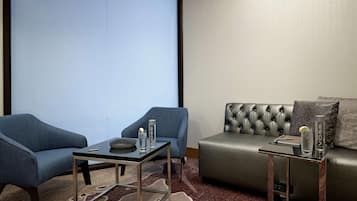 Ett modernt hotellrum med en grå soffa, två blå fåtöljer, ett glasbord och ett litet sidobord med en flaska och ett glas.