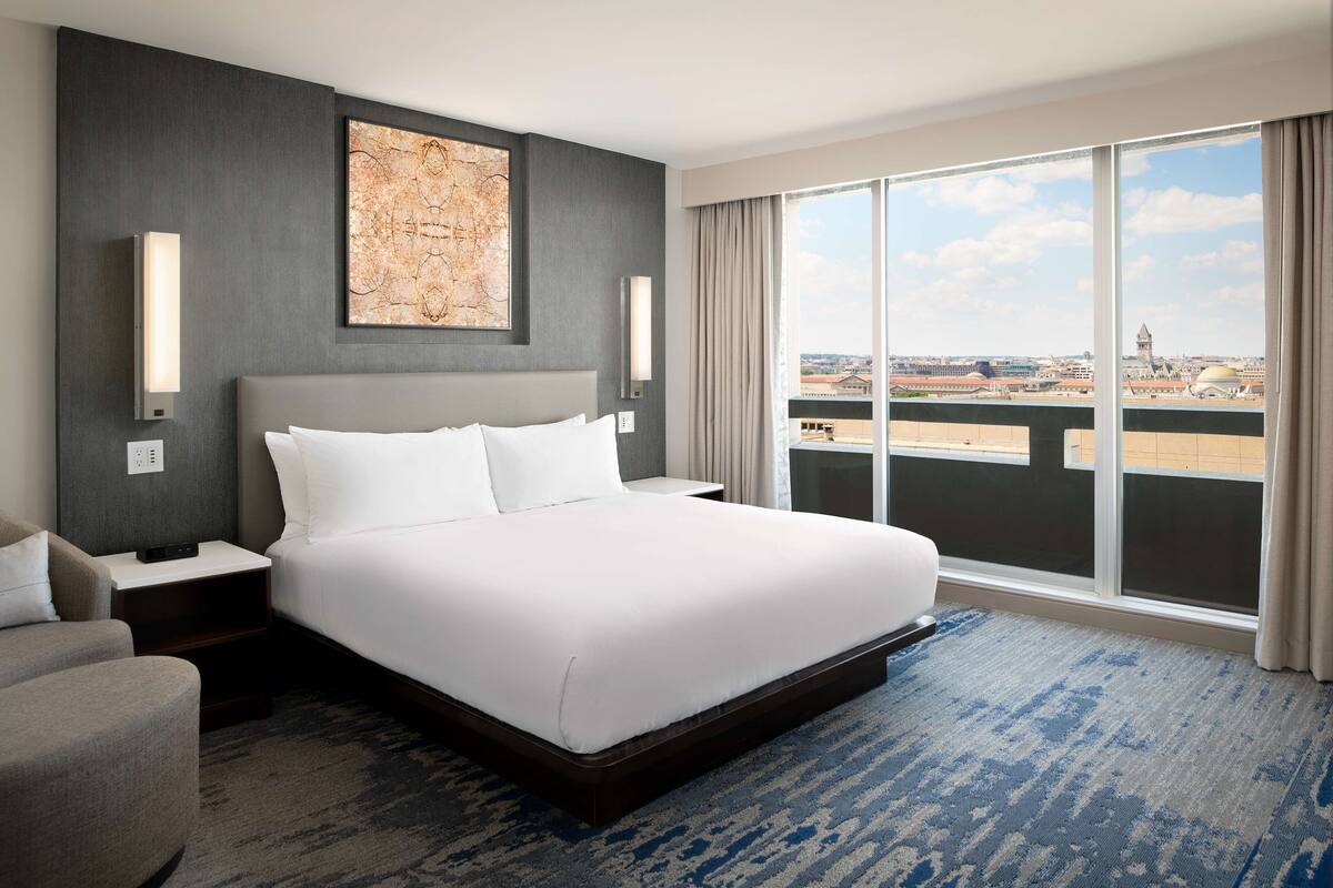 A hotel room with a large bed, a nightstand, a chair, a large window with a city view, and a wall-mounted artwork.