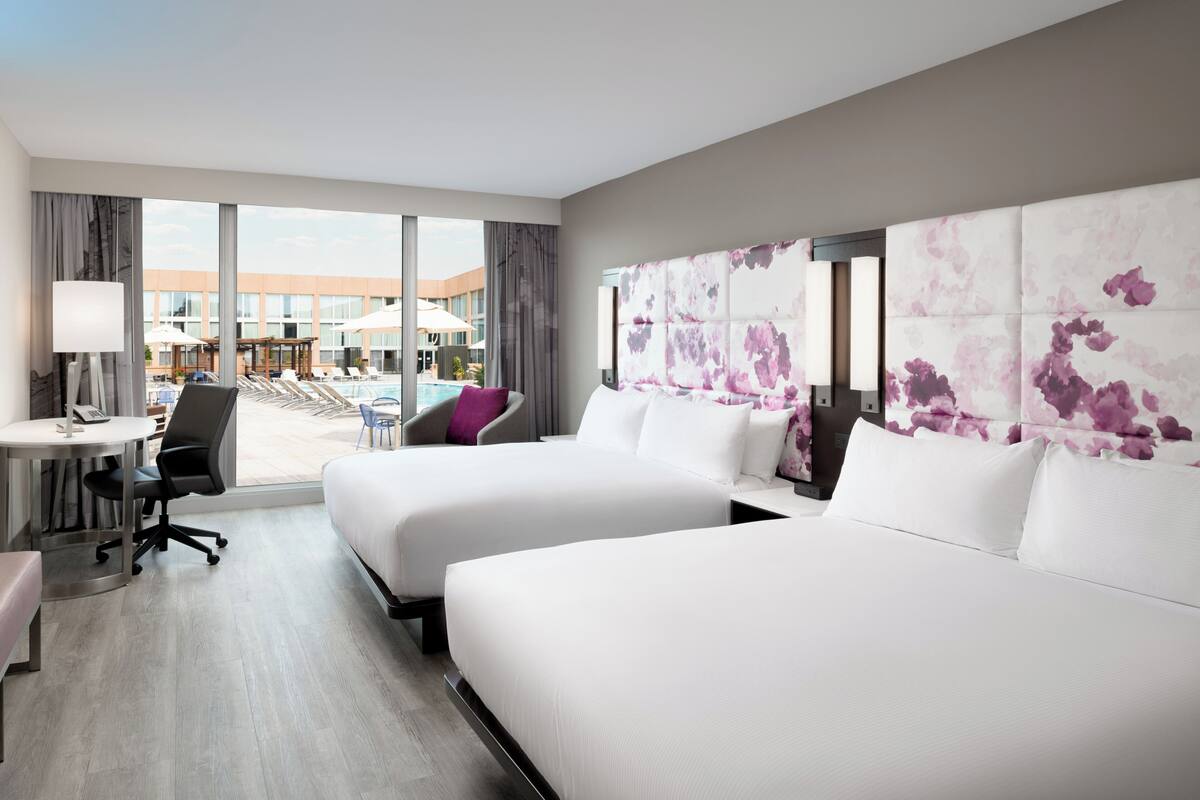 A hotel room with two beds, a desk, a chair, and a view of the pool area.