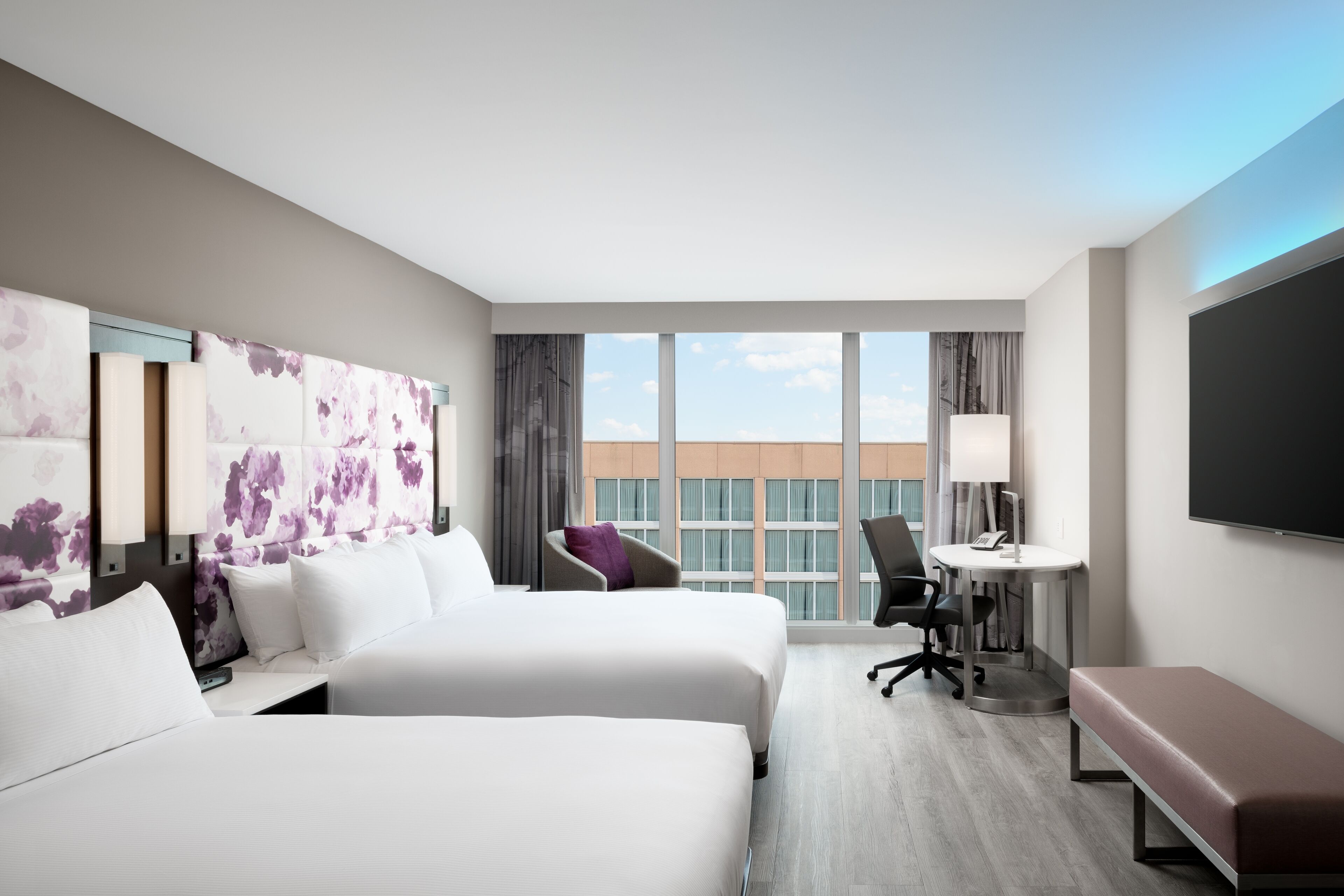Habitación de hotel con dos camas, un escritorio, una silla, un televisor y un ventanal con vistas a la ciudad.