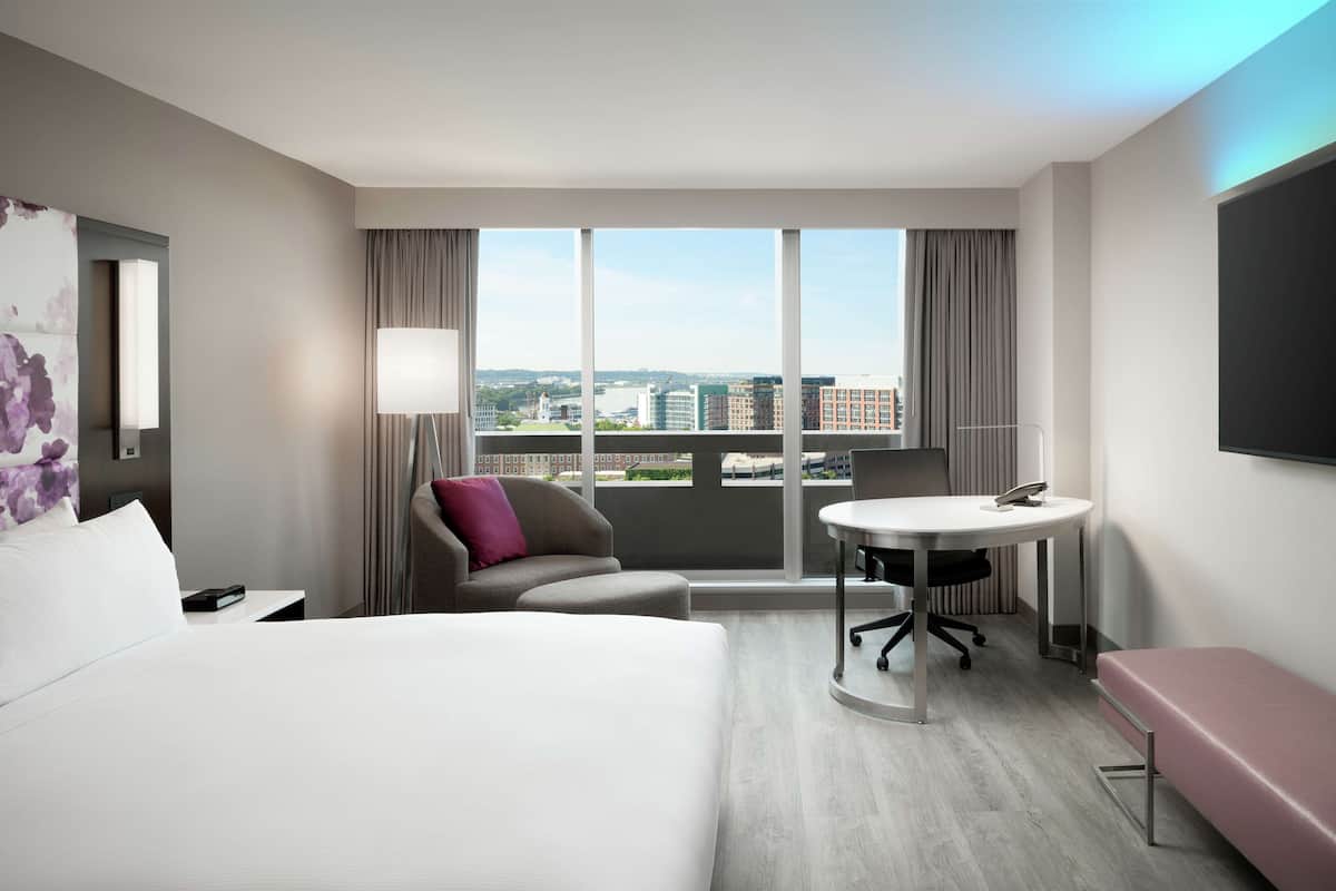A hotel room with a bed, a desk, a chair, a bench, a large window with a city view, and a flat-screen TV.