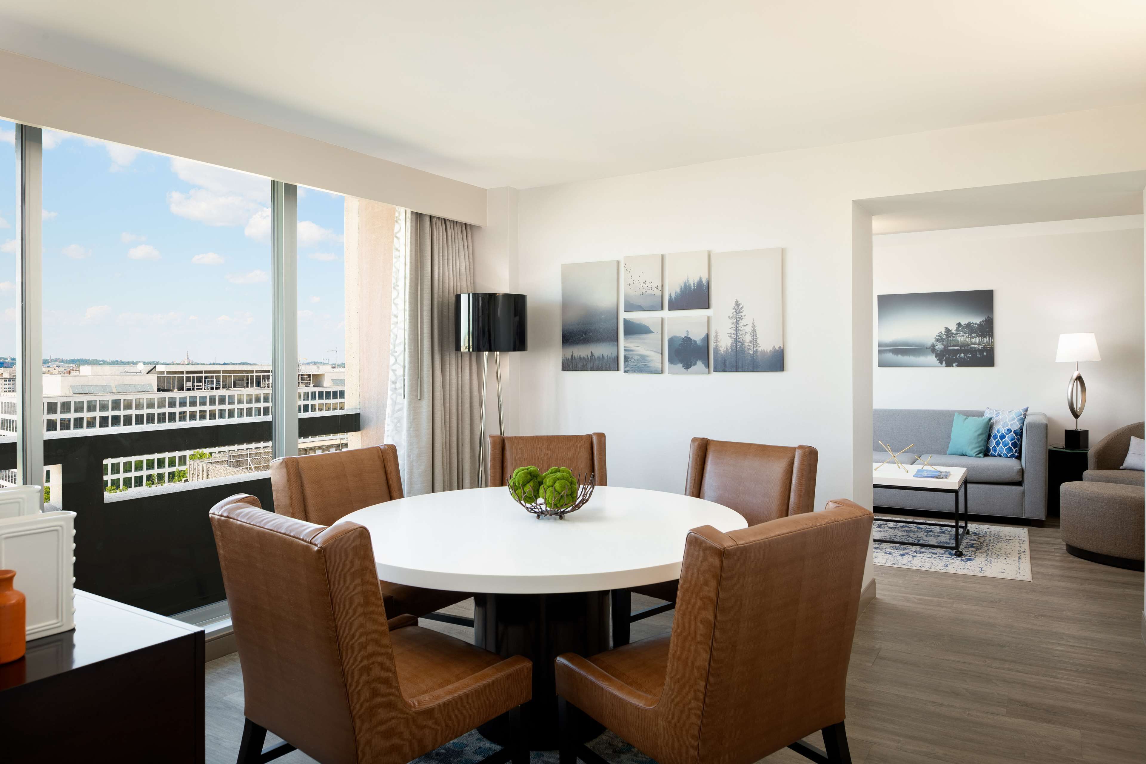 A modern hotel room with a round dining table, brown leather chairs, a flat-screen TV on the wall, and a large window offering a city view.