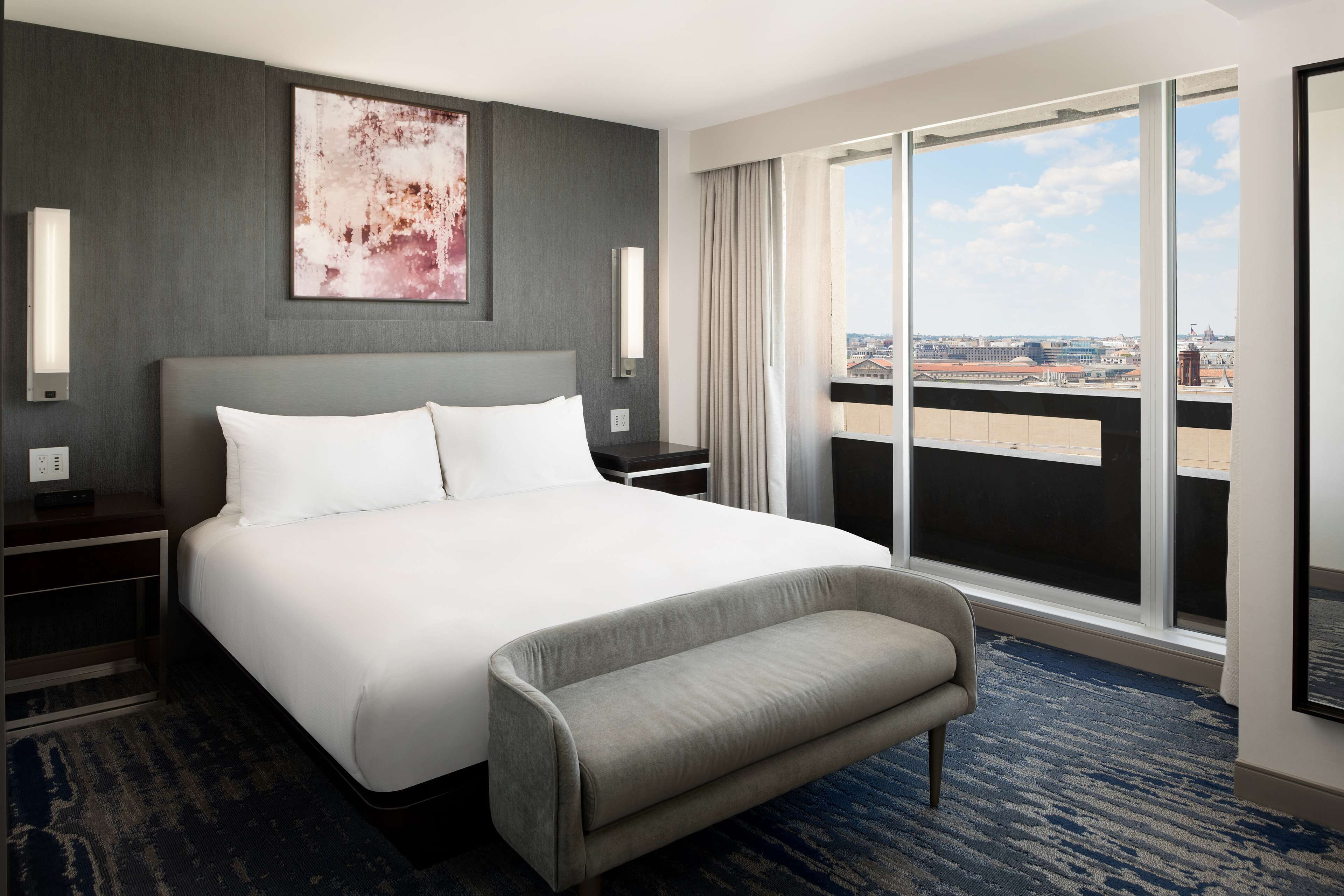 A hotel room with a large bed, a bench, a city view through the window, and a painting on the wall.