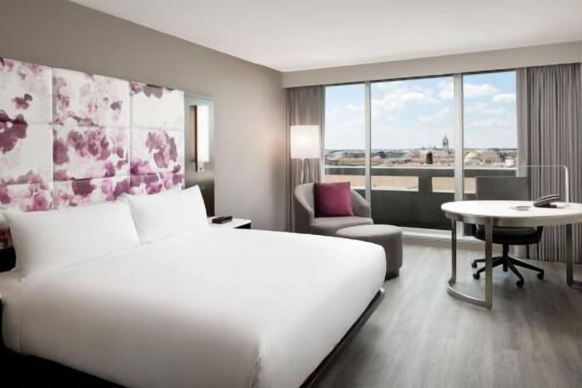 A hotel room with a large bed, a desk with a chair, a view of the cityscape, and a window with curtains.
