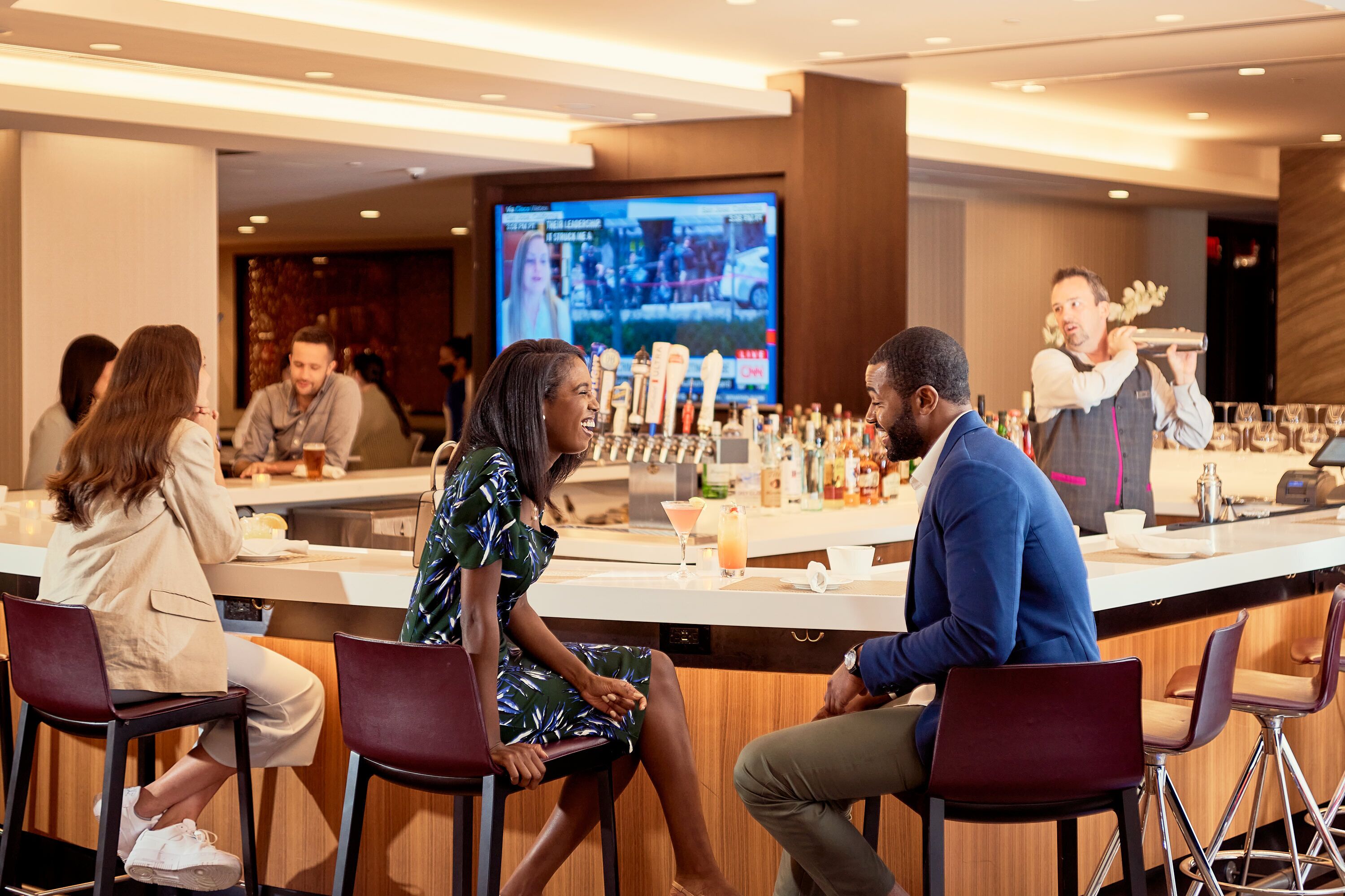 A bar area with stools, a bartender preparing drinks, and a television screen in the background.