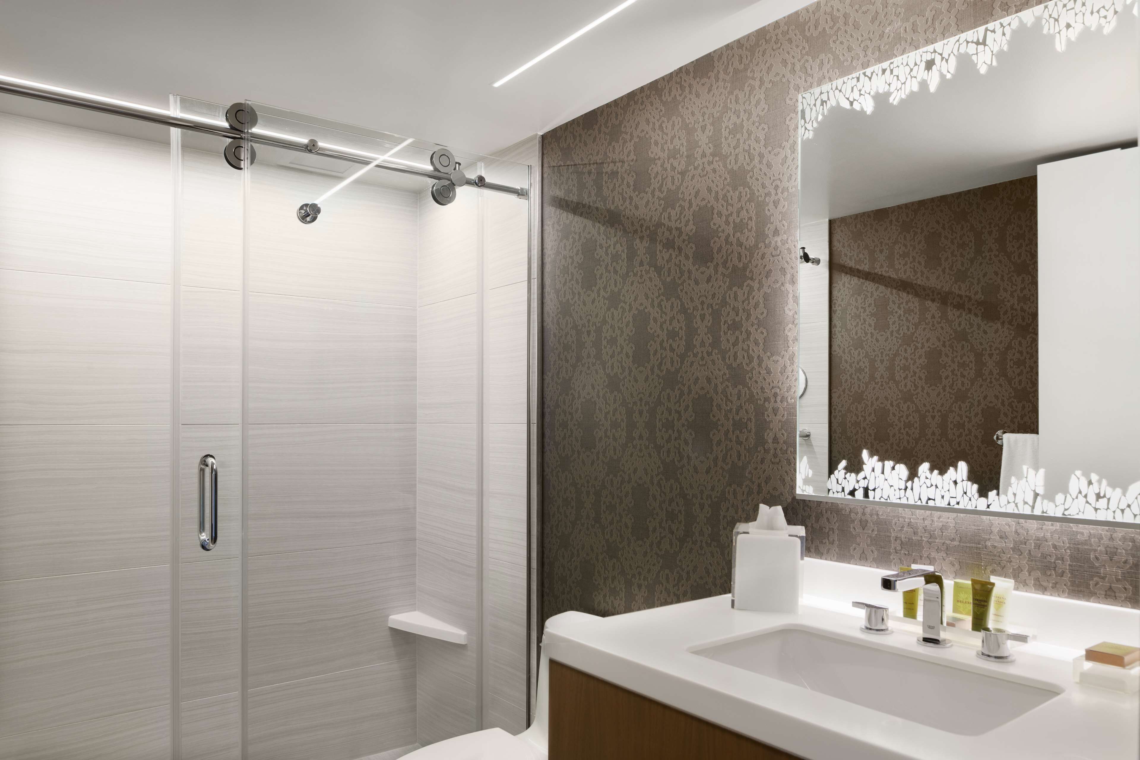 A modern bathroom with a glass shower enclosure, a white sink with a mirror, and a patterned wall.