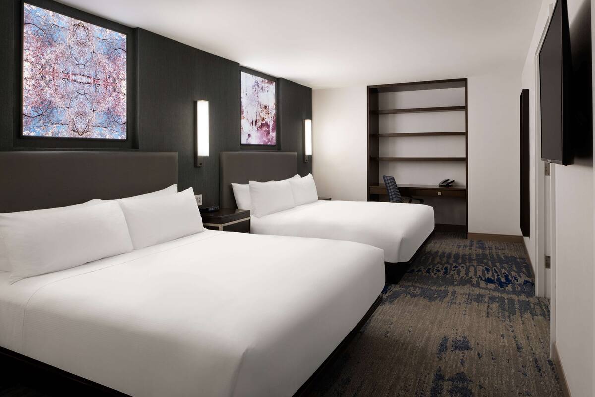 A hotel room with two beds, a desk, a chair, and a bookshelf.