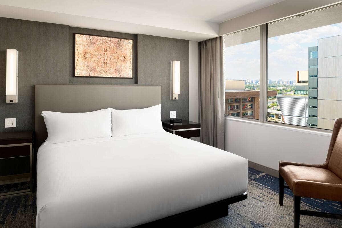 A hotel room with a large bed, a chair, a nightstand, a wall-mounted lamp, and a window with a city view.