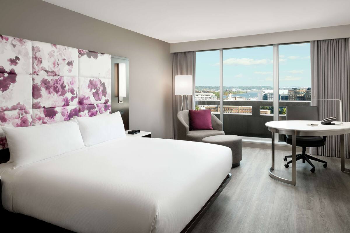 A hotel room with a large bed, a desk with a chair, a view of the city through large windows, and a decorative wall panel.