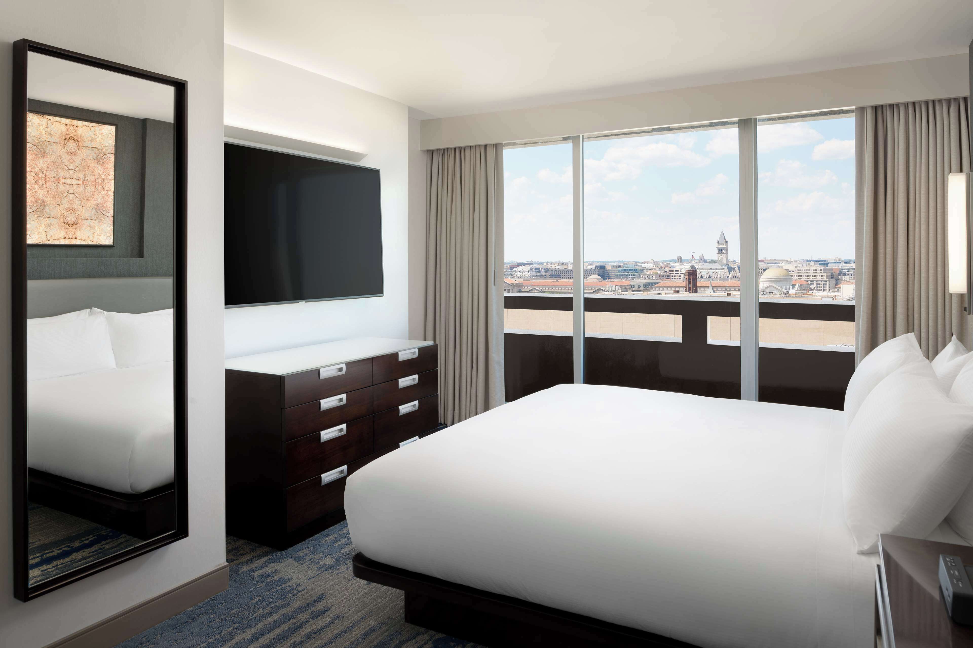 A hotel room with a large bed, a flat-screen TV, a dresser, and a city view through large windows.