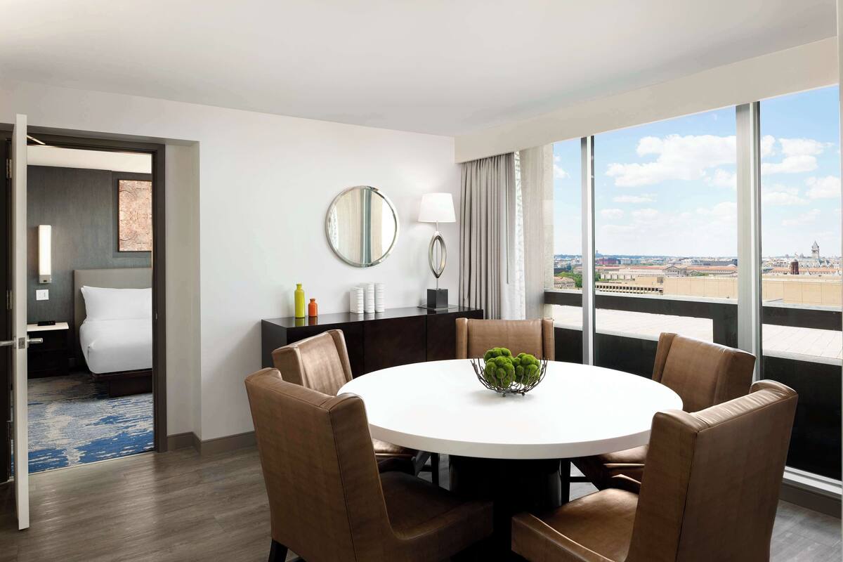 A hotel room with a round dining table, brown upholstered chairs, a round mirror, a lamp, and a view of the city through large windows.
