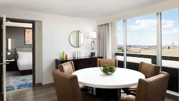 A hotel room with a round dining table, brown upholstered chairs, a round mirror, a lamp, and a view of the city through large windows.