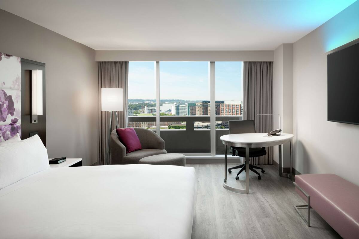 A hotel room with a large bed, a desk with a chair, a small sofa, a bench, and a large window offering a city view.
