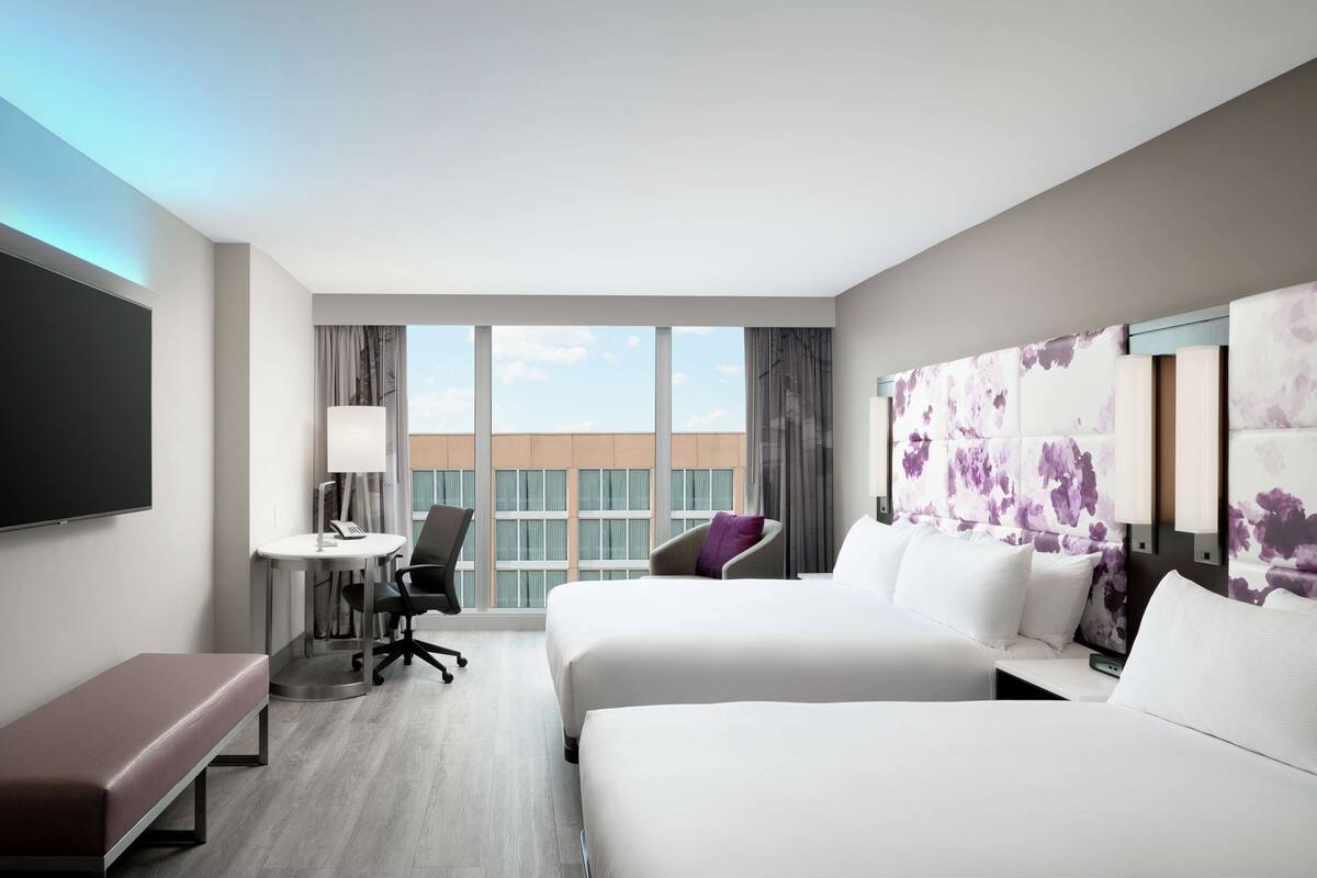 A hotel room with two beds, a desk, a chair, a bench, and a large window offering a view of buildings.