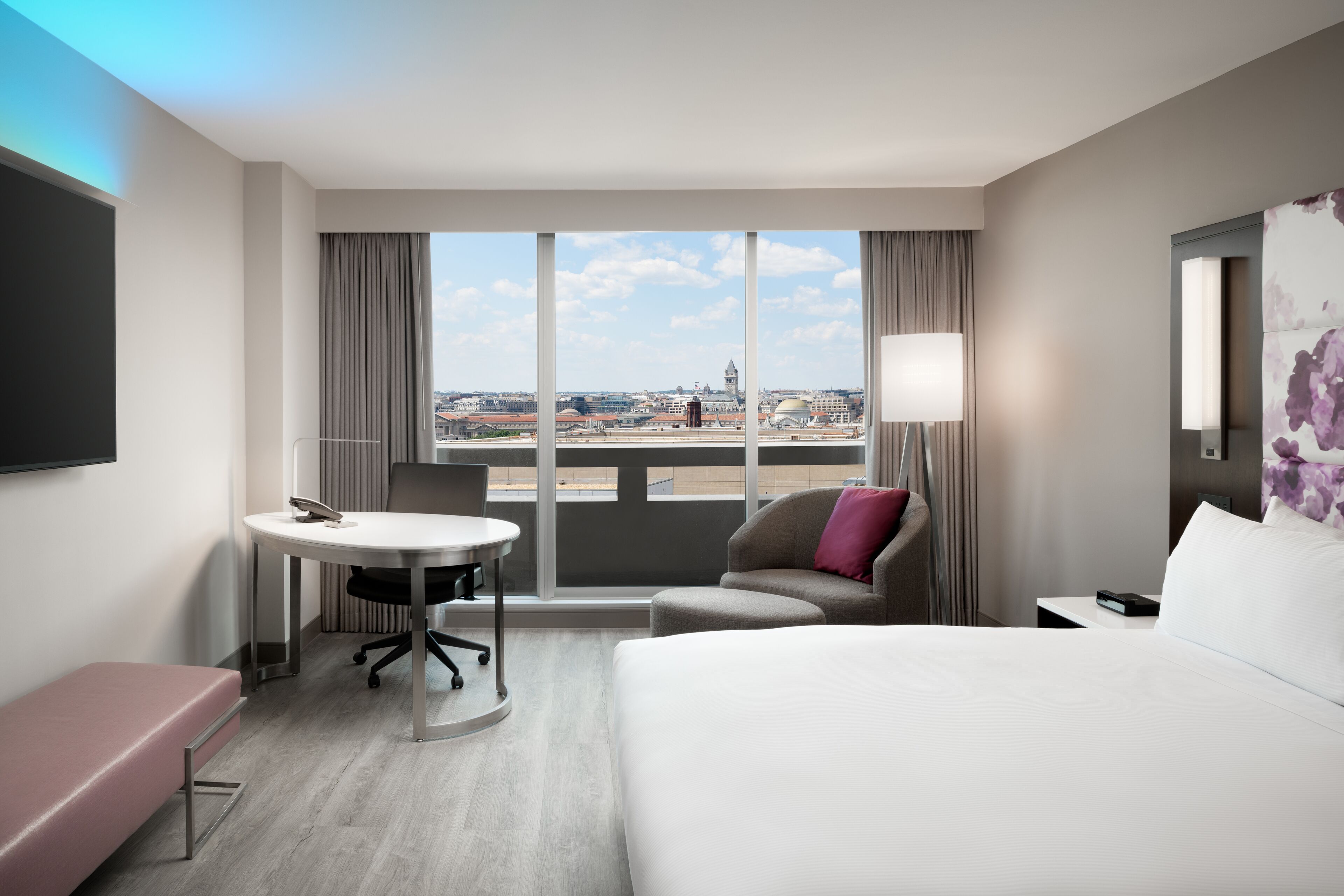 A hotel room with a large bed, a desk, a chair, and a view of the cityscape through large windows.