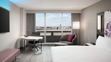 A hotel room with a large bed, a desk, a chair, and a view of the cityscape through large windows.