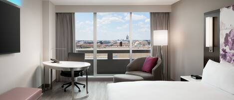 A hotel room with a large bed, a desk, a chair, and a view of the cityscape through large windows.