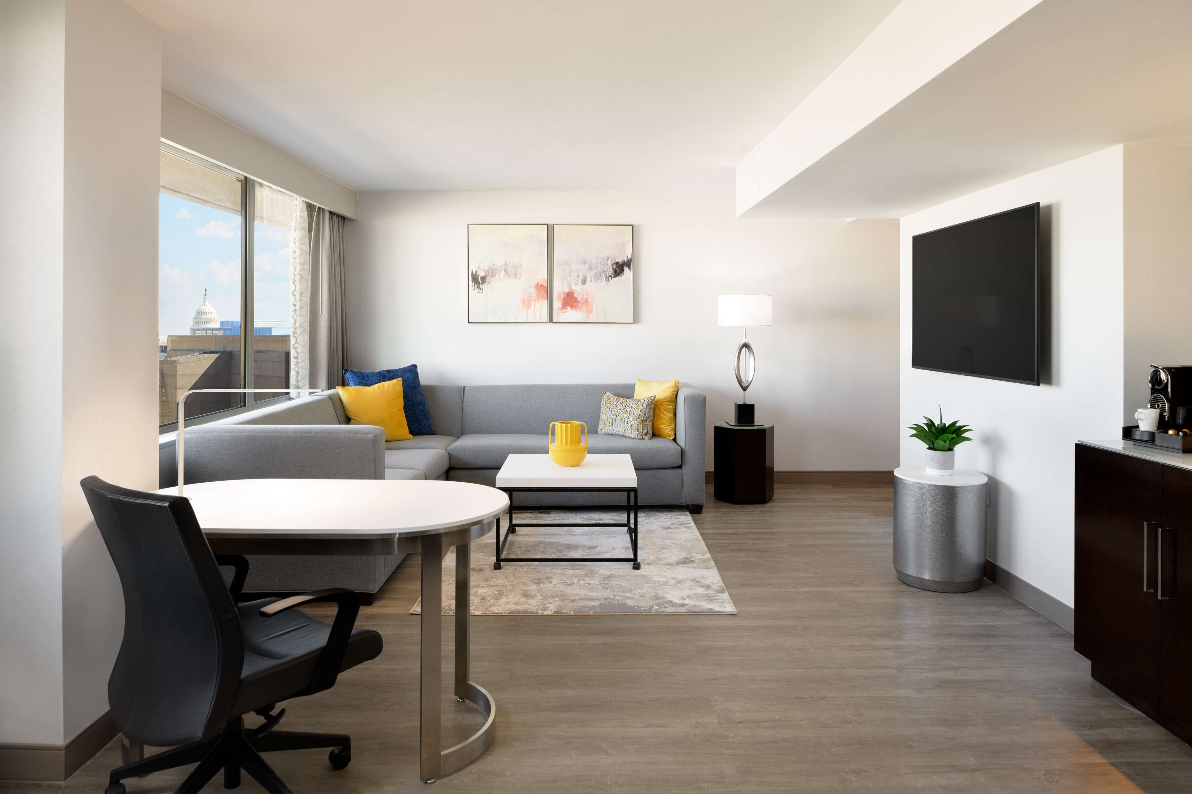 A modern living room with a gray sofa, a coffee table, a dining table with chairs, a TV mounted on the wall, and a large window with a view of a building.