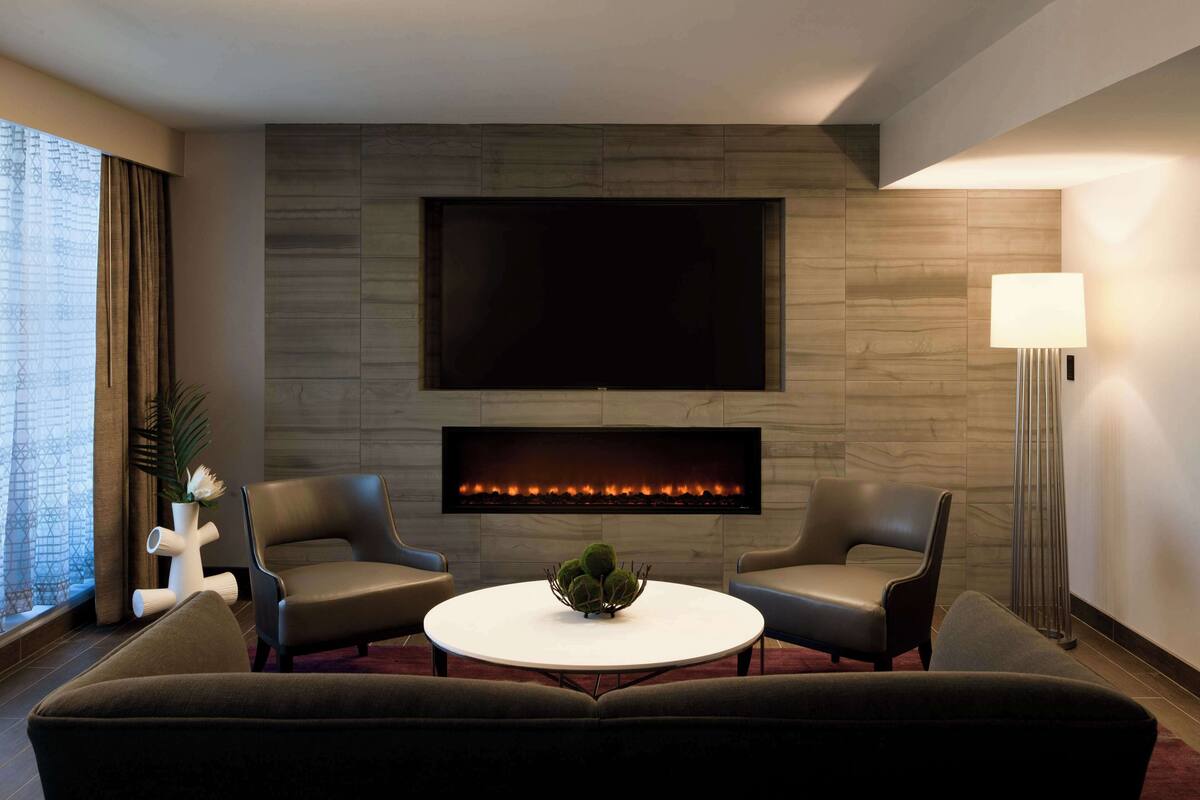 A modern living room with a flat-screen TV, a fireplace, two leather armchairs, a round coffee table with a green centerpiece, and a floor lamp.