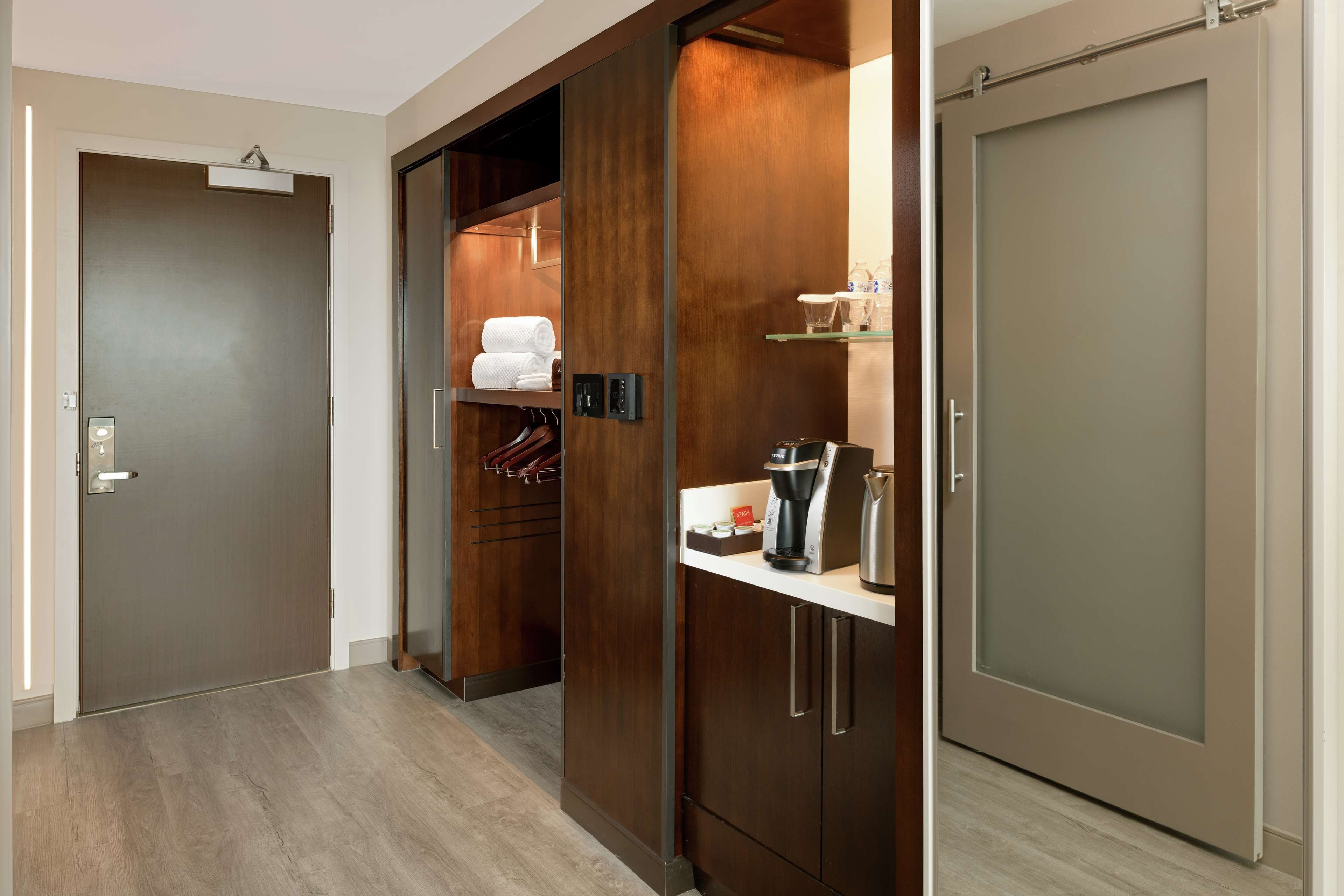 A hotel room with a wooden wardrobe, a coffee machine, and a shelf with toiletries.