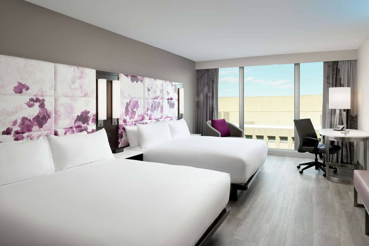 A hotel room with two beds, a desk, a chair, and a view of the city.
