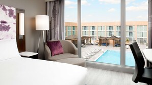 A hotel room with a large window, a bed, a sofa, a lamp, and a view of a pool and outdoor seating area. - Hilton Washington DC National Mall The Wharf (Washington)
