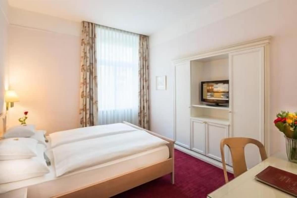 Habitación de hotel con cama, televisor sobre un mueble, escritorio con silla y ventana con cortinas.