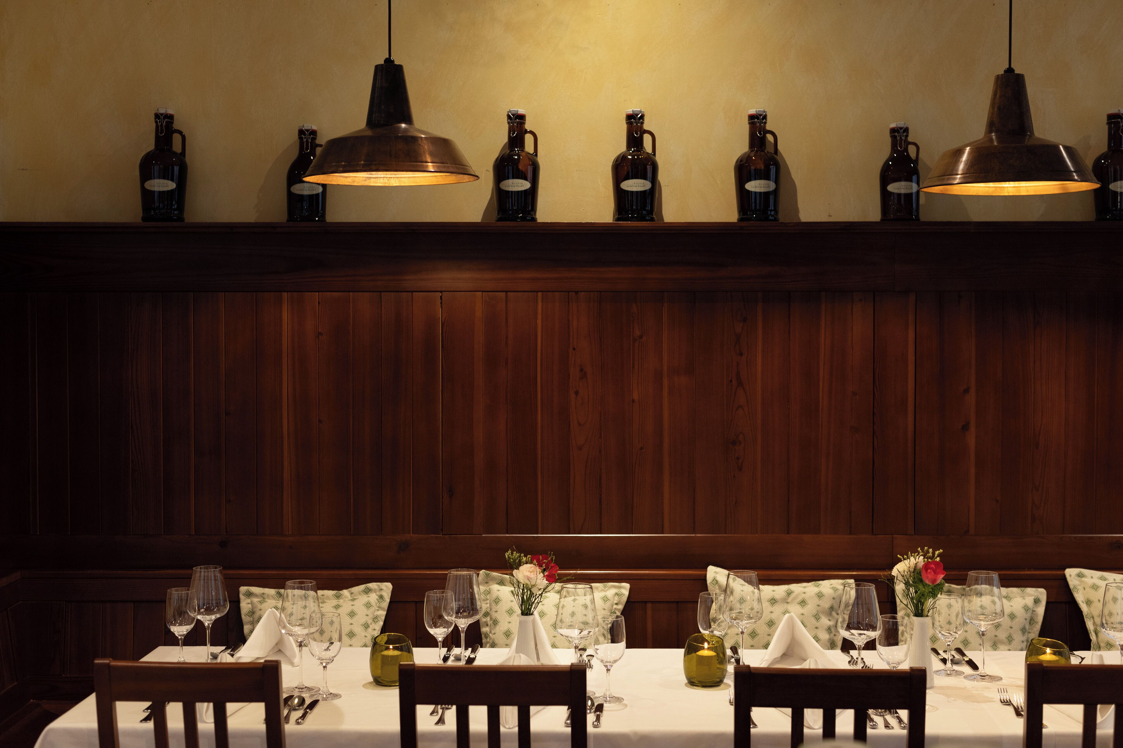 Un restaurant aux murs lambrés, une longue table dressée avec des verres à vin et des bouteilles exposées sur le mur.