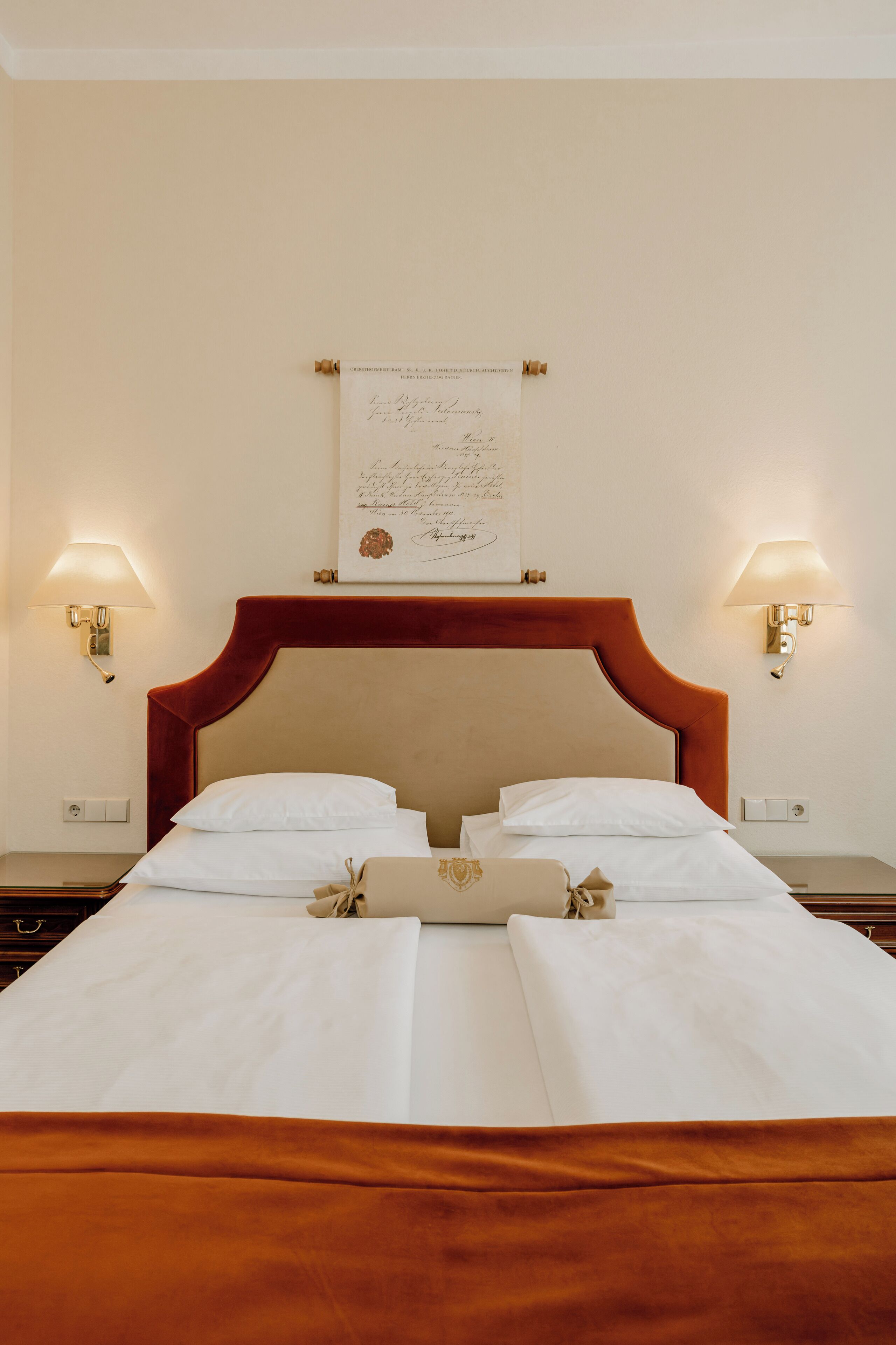 A neatly made bed with white linens and a brown headboard, two bedside lamps, and a framed document on the wall.