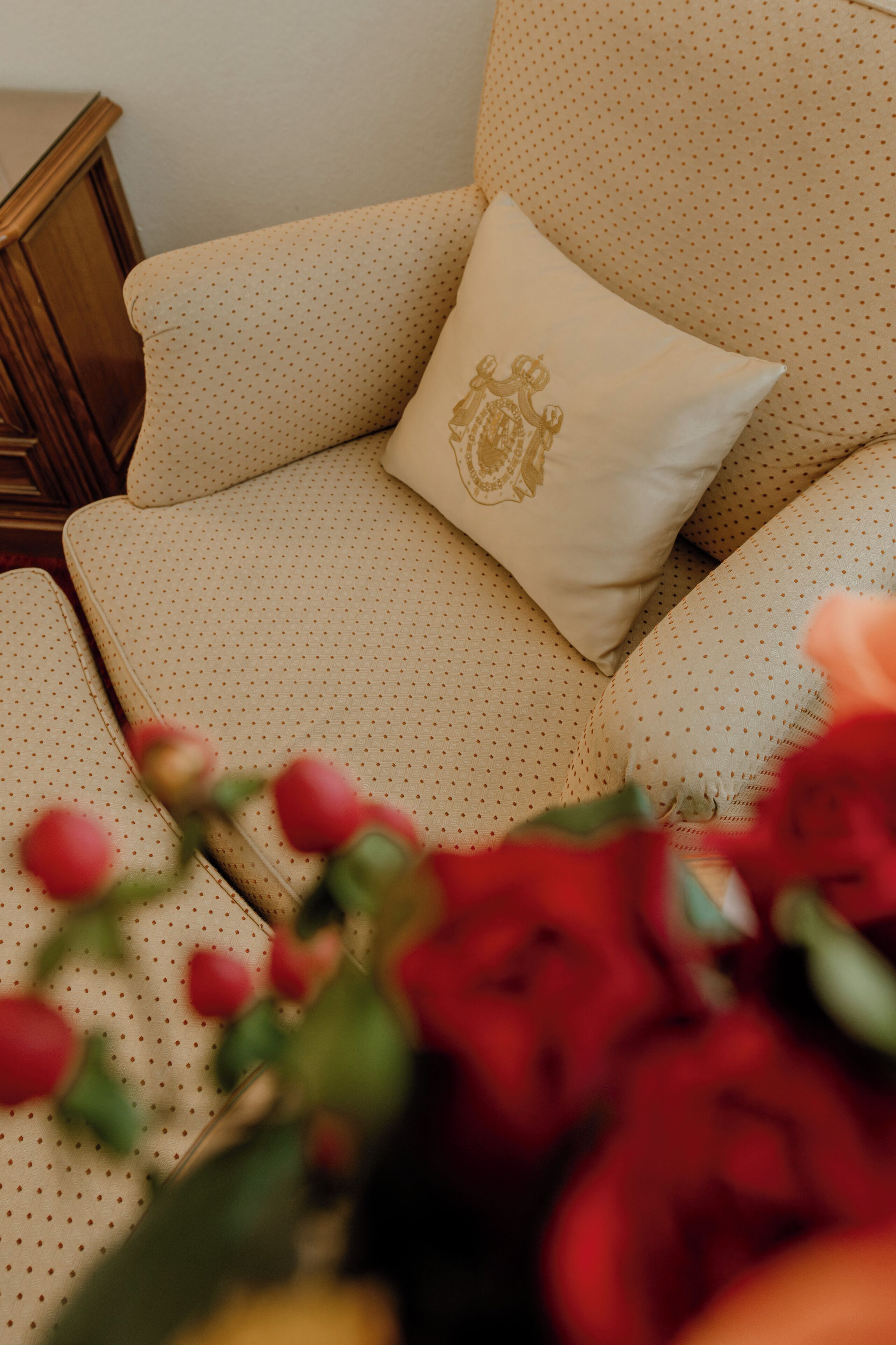 A beige sofa with a gold embroidered pillow, a wooden nightstand, and red flowers in the foreground.