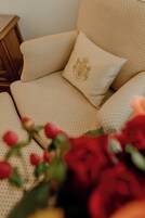 Um sofá bege com um travesseiro bordado dourado, um criado-mudo de madeira e flores vermelhas em primeiro plano.