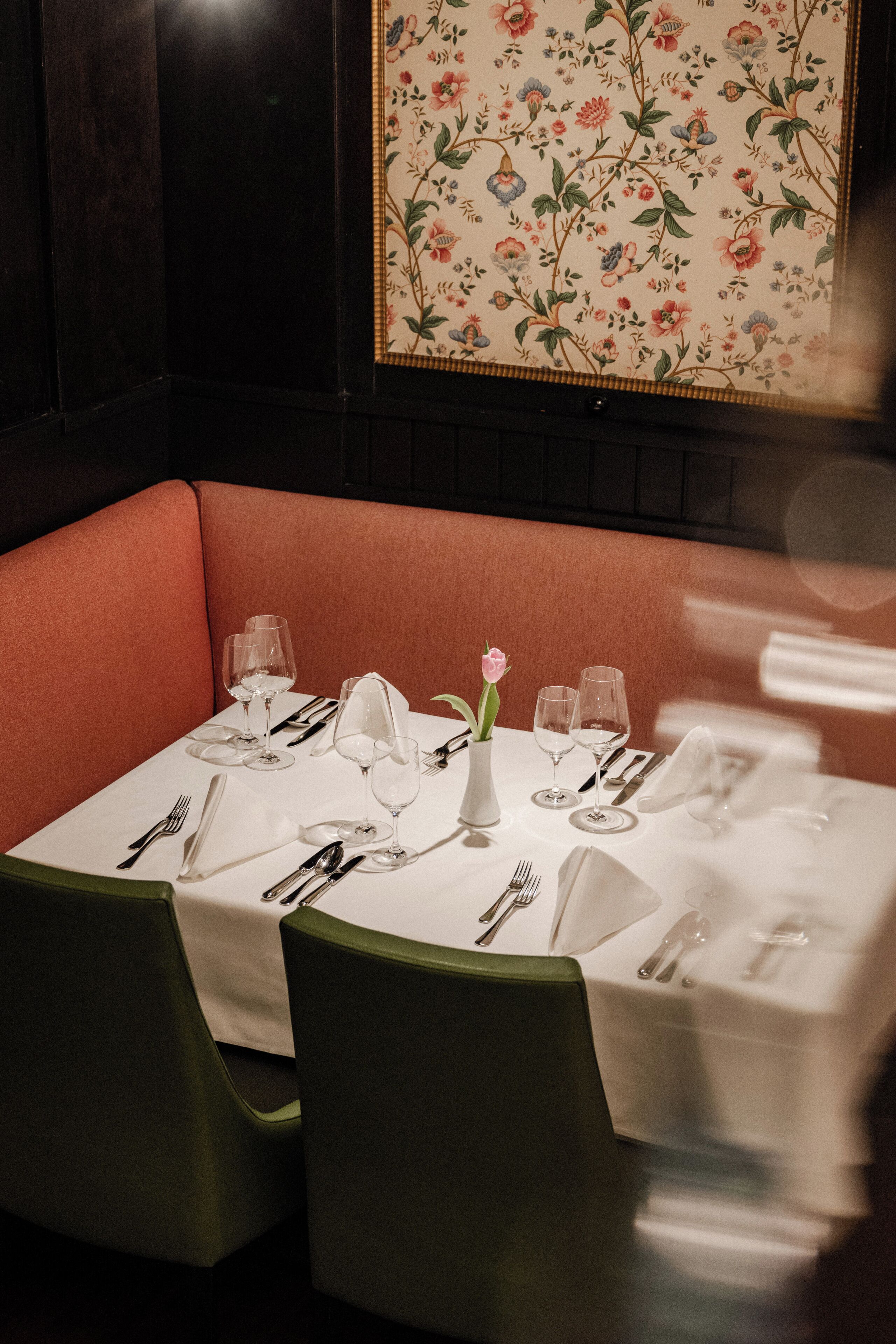 A dining table set with white tablecloth, silverware, and wine glasses, surrounded by green upholstered chairs.