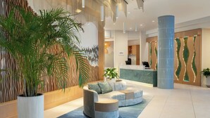 A modern lobby with a reception desk, a large potted plant, a sectional sofa, and a decorative wall.
