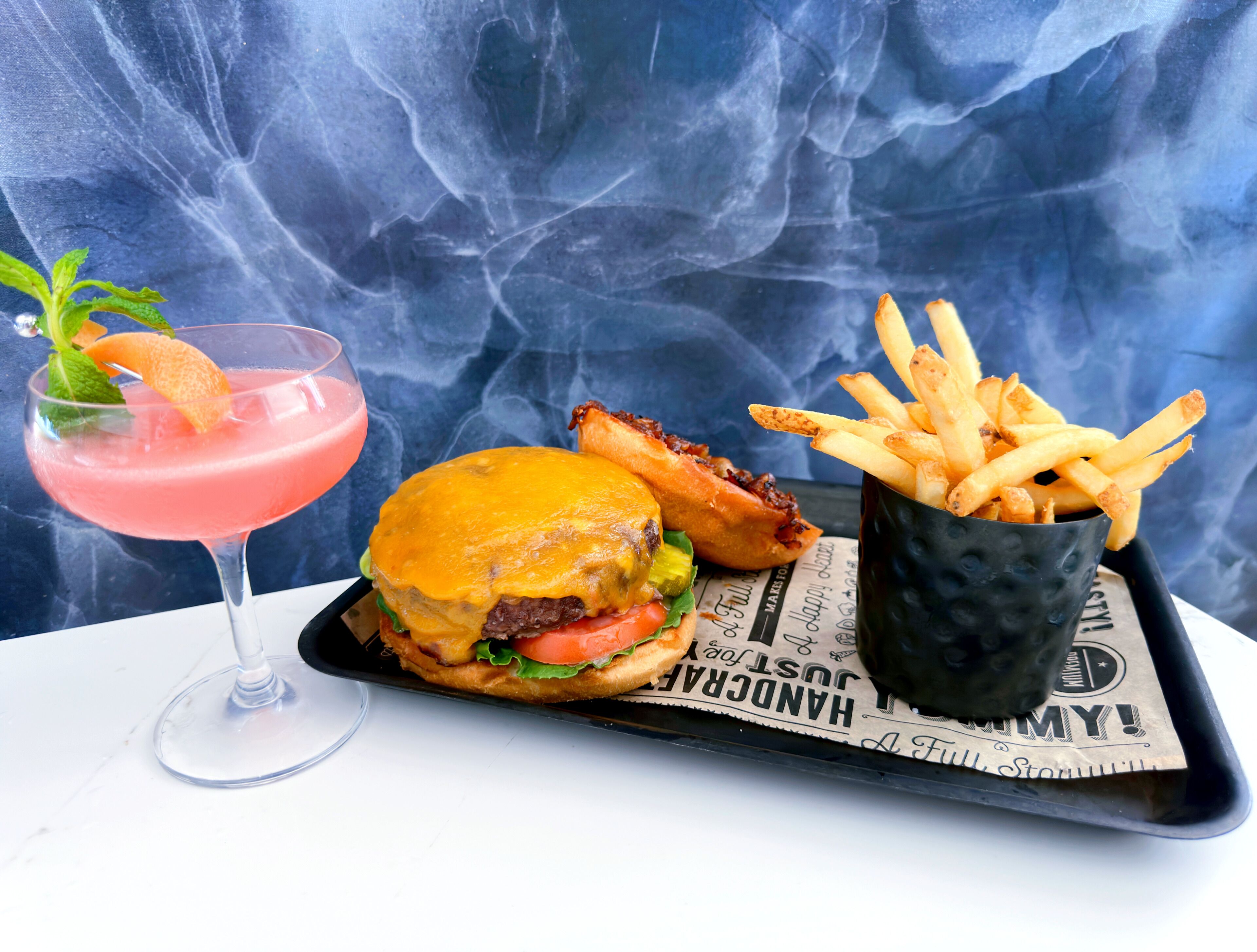 A burger, fries, and a pink drink on a tray with a newspaper underneath.