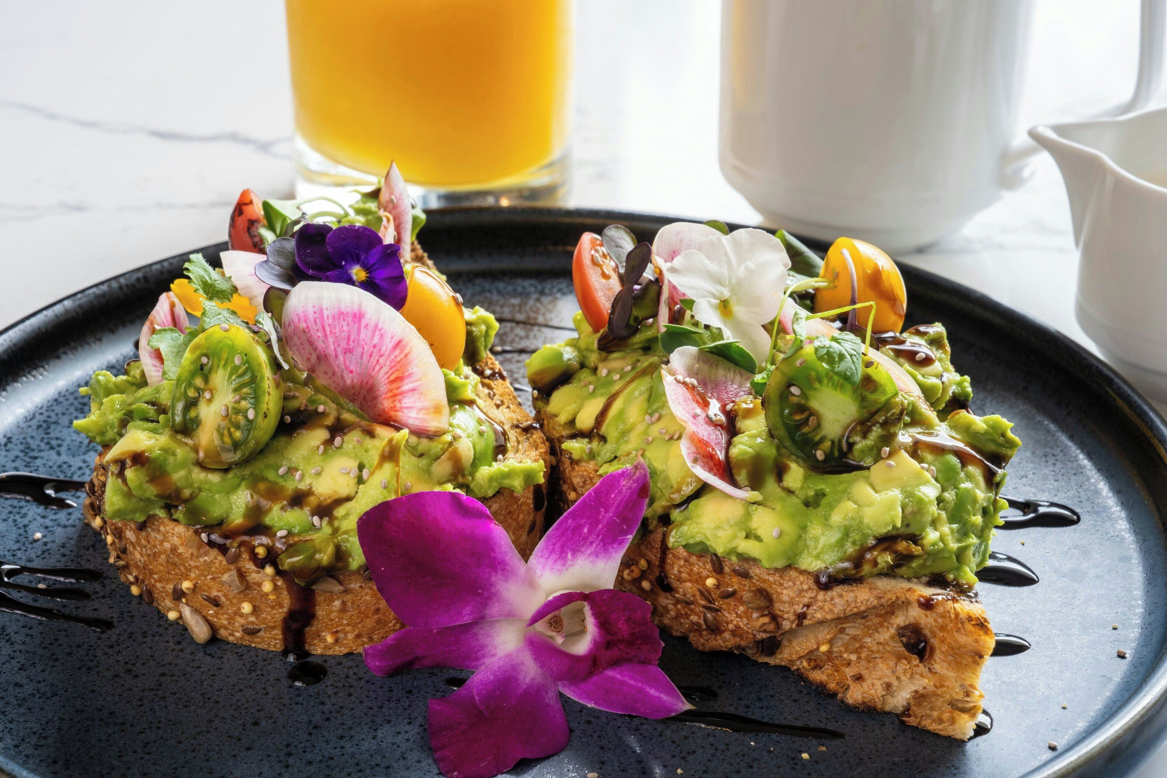 Two avocado toast sandwiches on a plate with a purple orchid.