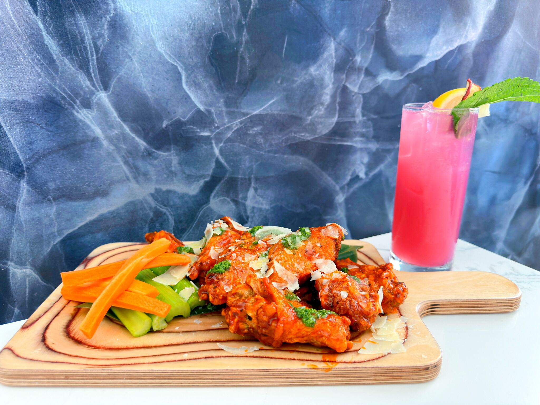 A meal with chicken wings, vegetables, and a pink drink on a wooden board.