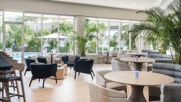 A spacious indoor area with multiple seating arrangements, including sofas and chairs, near large windows offering views of palm trees and a pool.