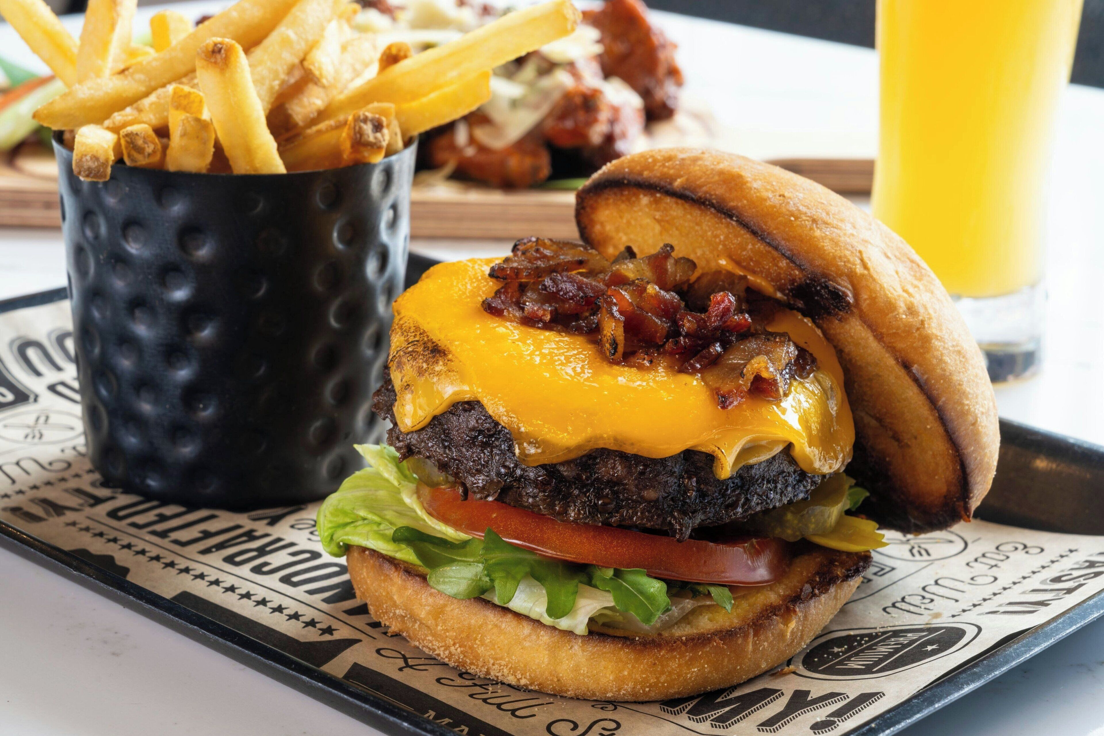 Una hamburguesa con tocino, queso, lechuga y tomate, servida junto a una porción de papas fritas y un vaso de jugo de naranja.
