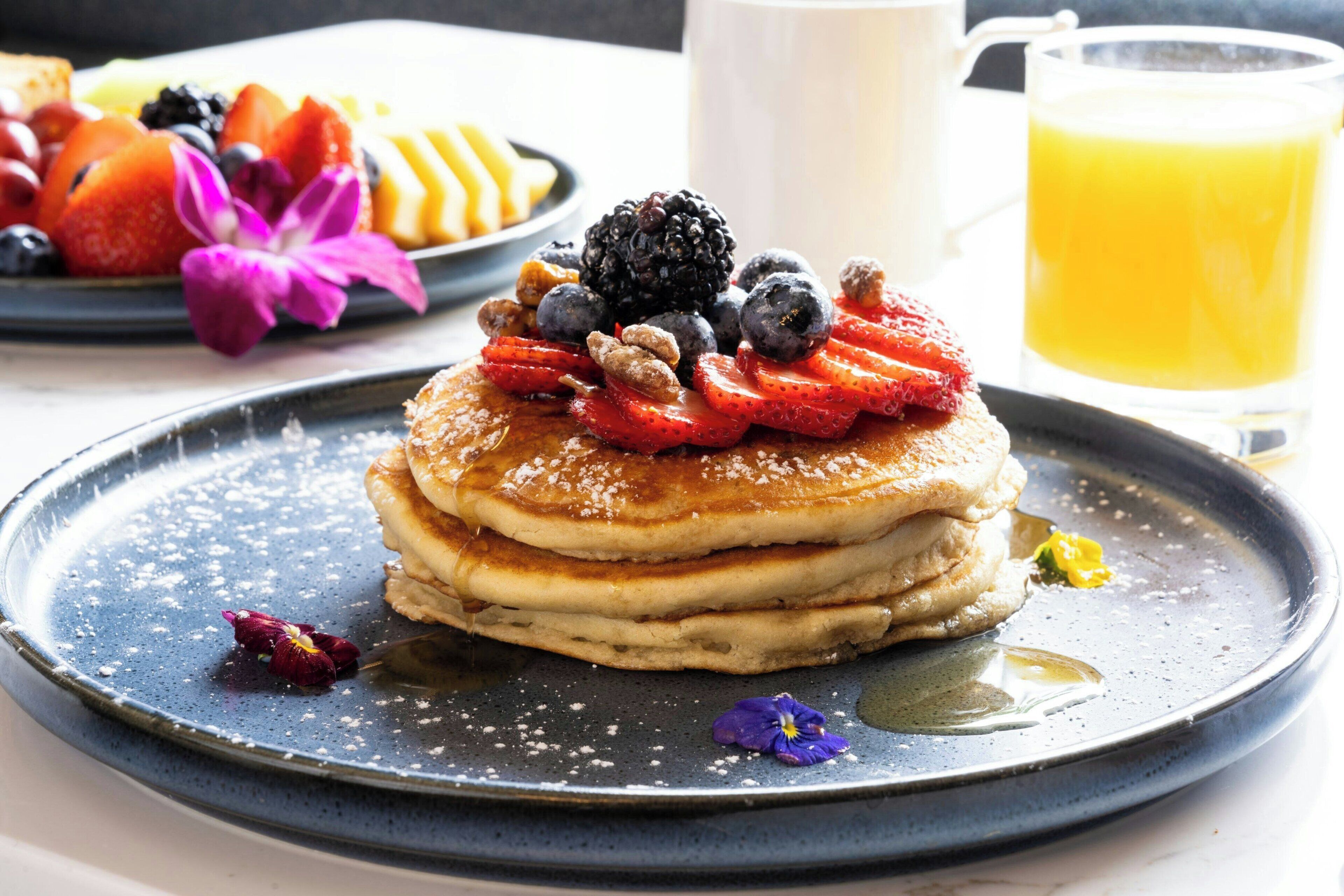 Une pile de crêpes garnie de fraises, de myrtilles et de mûres, servie sur une assiette bleue avec du sirop et décorée d'une fleur.