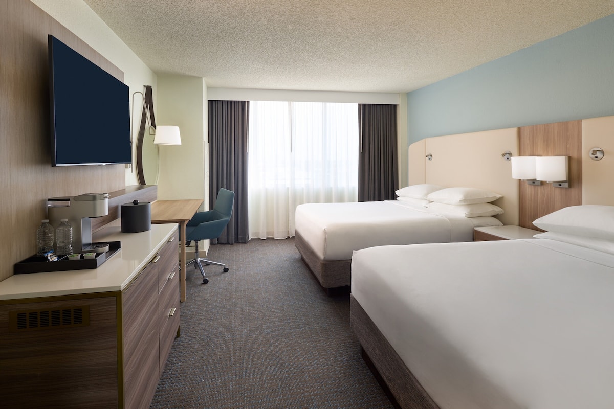 A hotel room with a flat-screen TV, a desk with a chair, two beds, a large window with curtains, and a wall-mounted lamp.