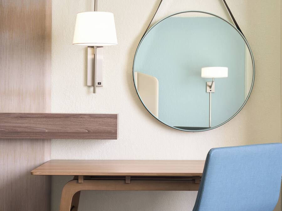 Une chambre d’hôtel moderne avec un bureau en bois, une chaise bleue, un grand miroir rond fixé au mur et deux appliques murales.