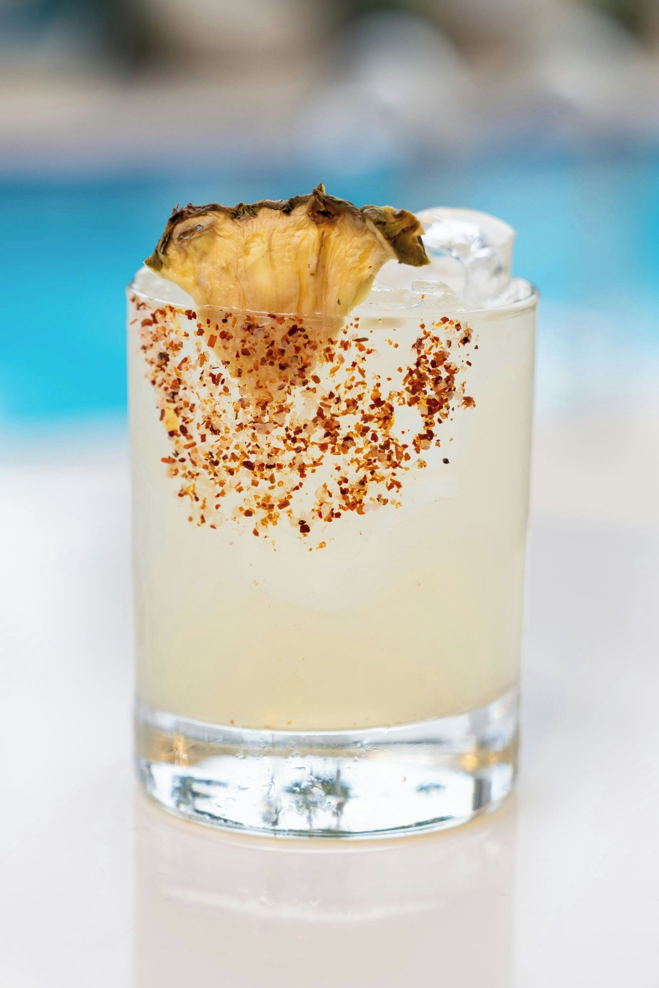 Un verre transparent contenant une boisson, des glaçons et une tranche d’ananas sur le bord, avec un arrière-plan flou.